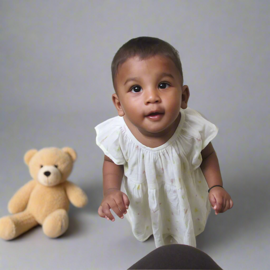 A girl baby wearing muslin frock and there is a teddy bear behind her 